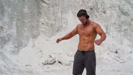 strong man with headphones is doing exercises on the mountain background. young shirtless male with muscular body is training in the open air in white mountains.