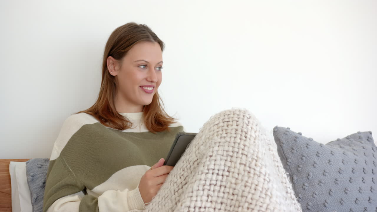 Relaxing on bed, young woman reading on tablet under cozy blanket