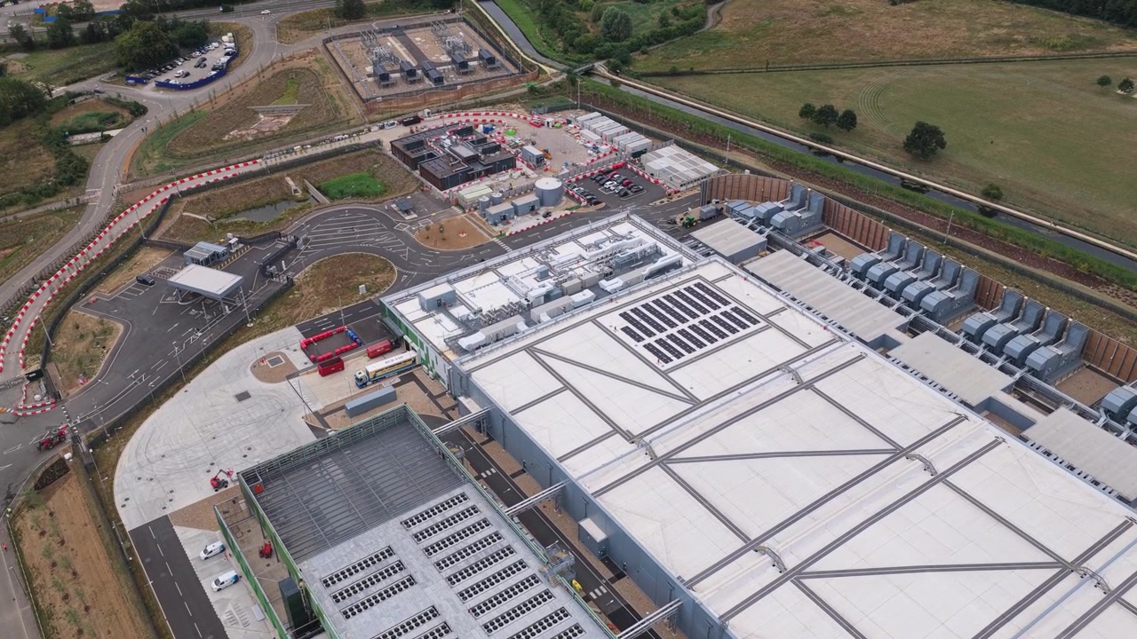 Stort industrielt datacenterkompleks omgivet af veje og grønne marker, fanget fra et forhøjet perspektiv i dagtimerne under en klar blå himmel i et landområde