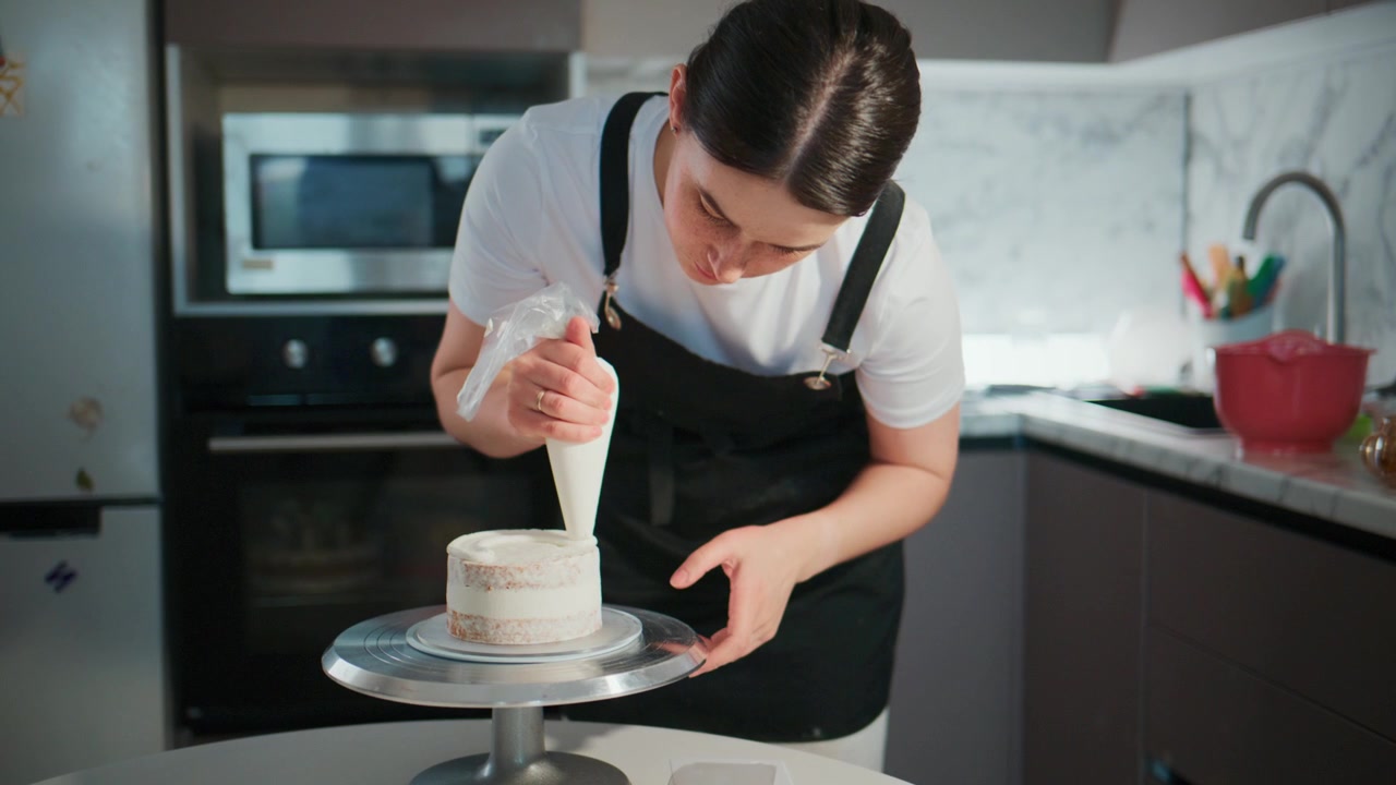 Fokusert baker sprøyter forsiktig glasur på en kake. Hun lener seg inn, konsentrerer seg med stødige hender. Kjøkkenet er rent og moderne, fylt med bakeutstyr. Ideell for oppskrifter, matlagingsprogrammer