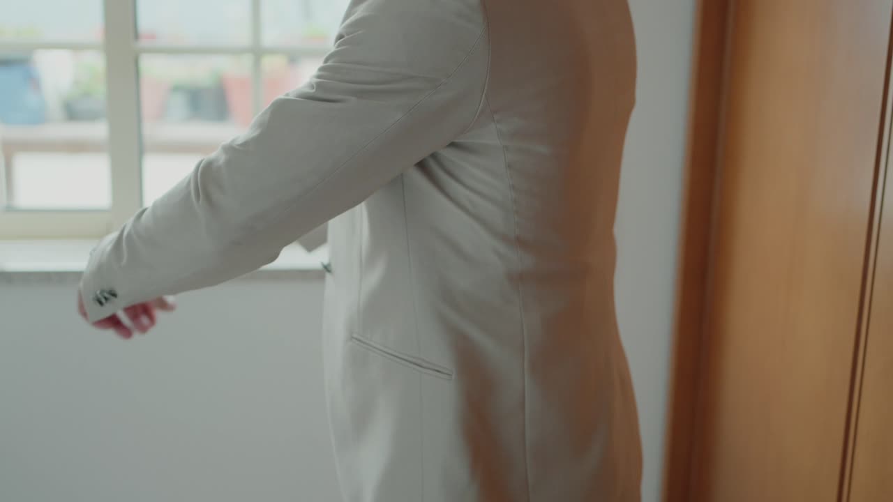 Groom putting on beige jacket near window with natural light and wooden interior