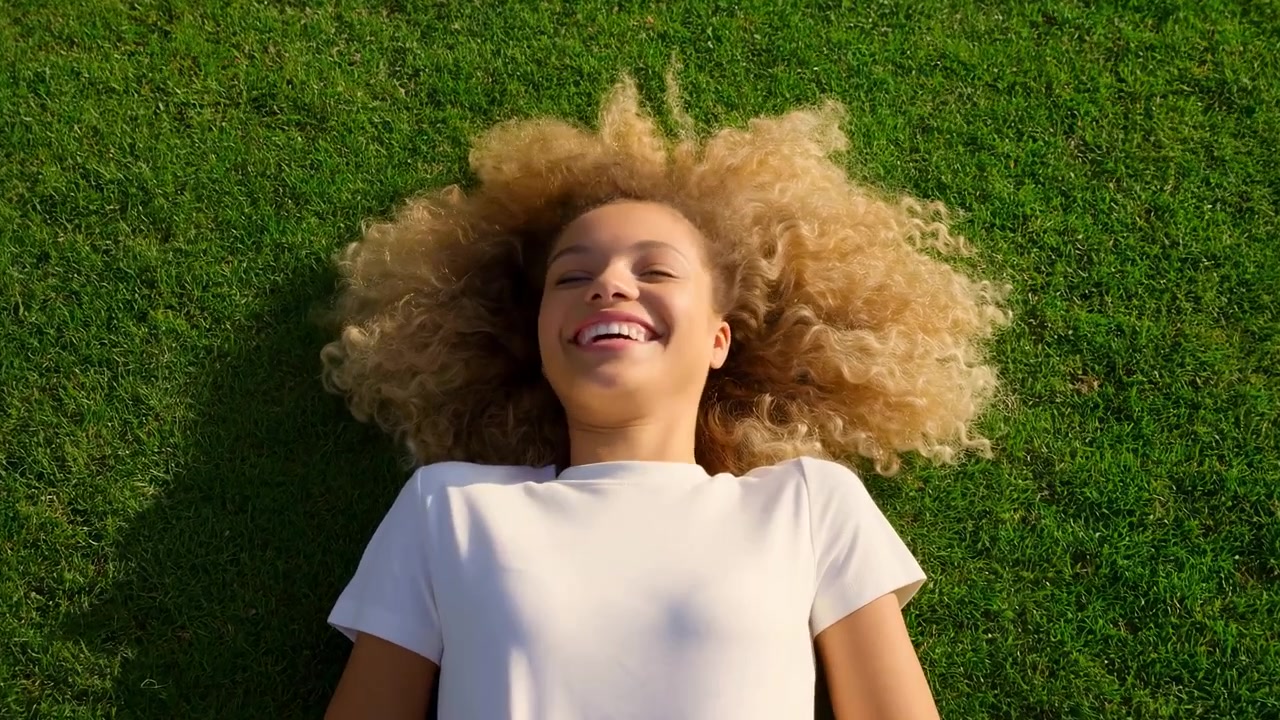 Biracial Woman Lying On Green Grass Smiling With Curly Blonde Hair Fanned Out Wearing White Tee Aerial View