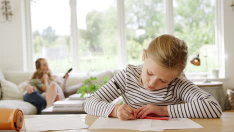 Girl-At-Table-Home-Schooling-During-Health-Pandemic-With-Family-In-Background