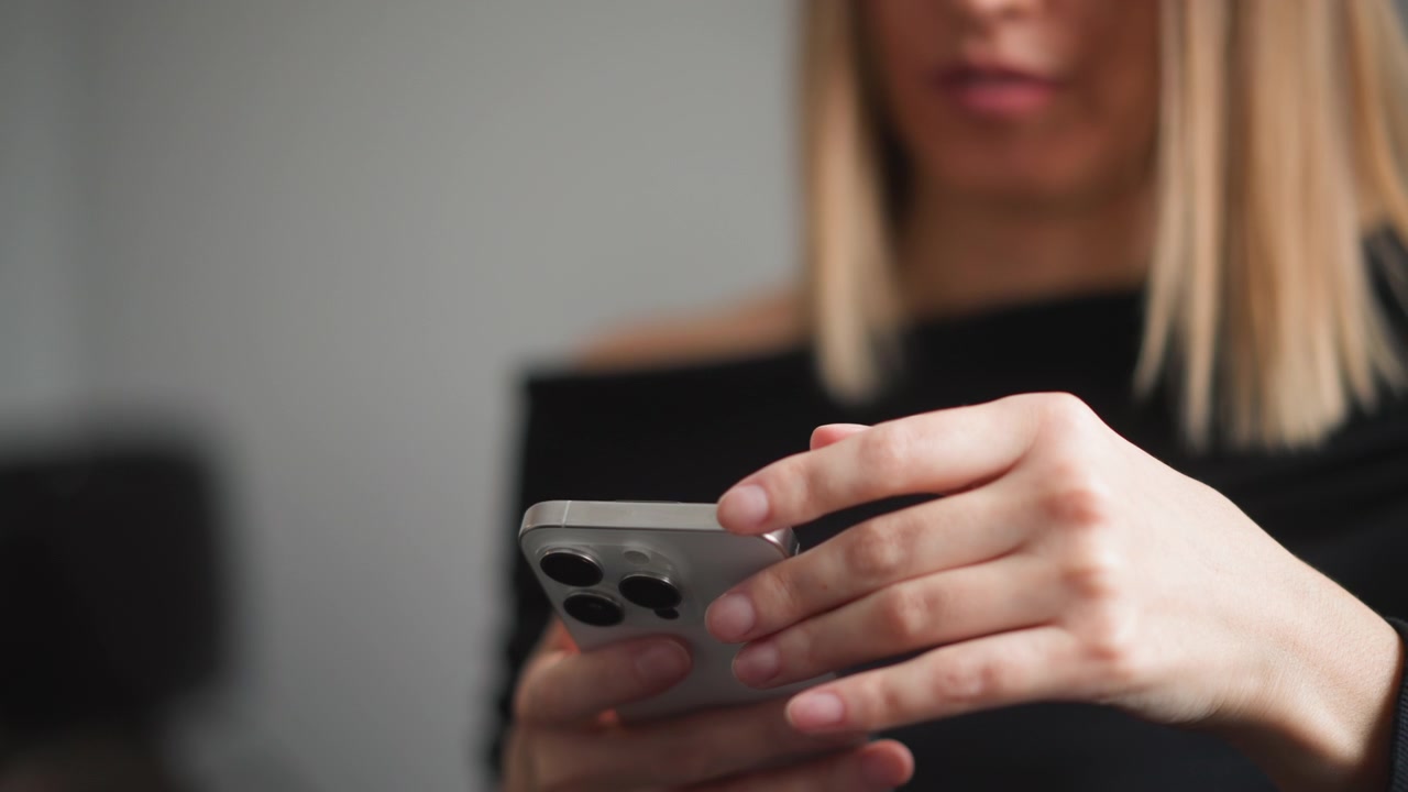 Primo piano di donna con smartphone tra le mani. La scena mostra le dita della donna che toccano o scorrono sul dispositivo, concentrandosi esclusivamente sui movimenti delle mani attorno al telefono. Illuminazione soffusa.