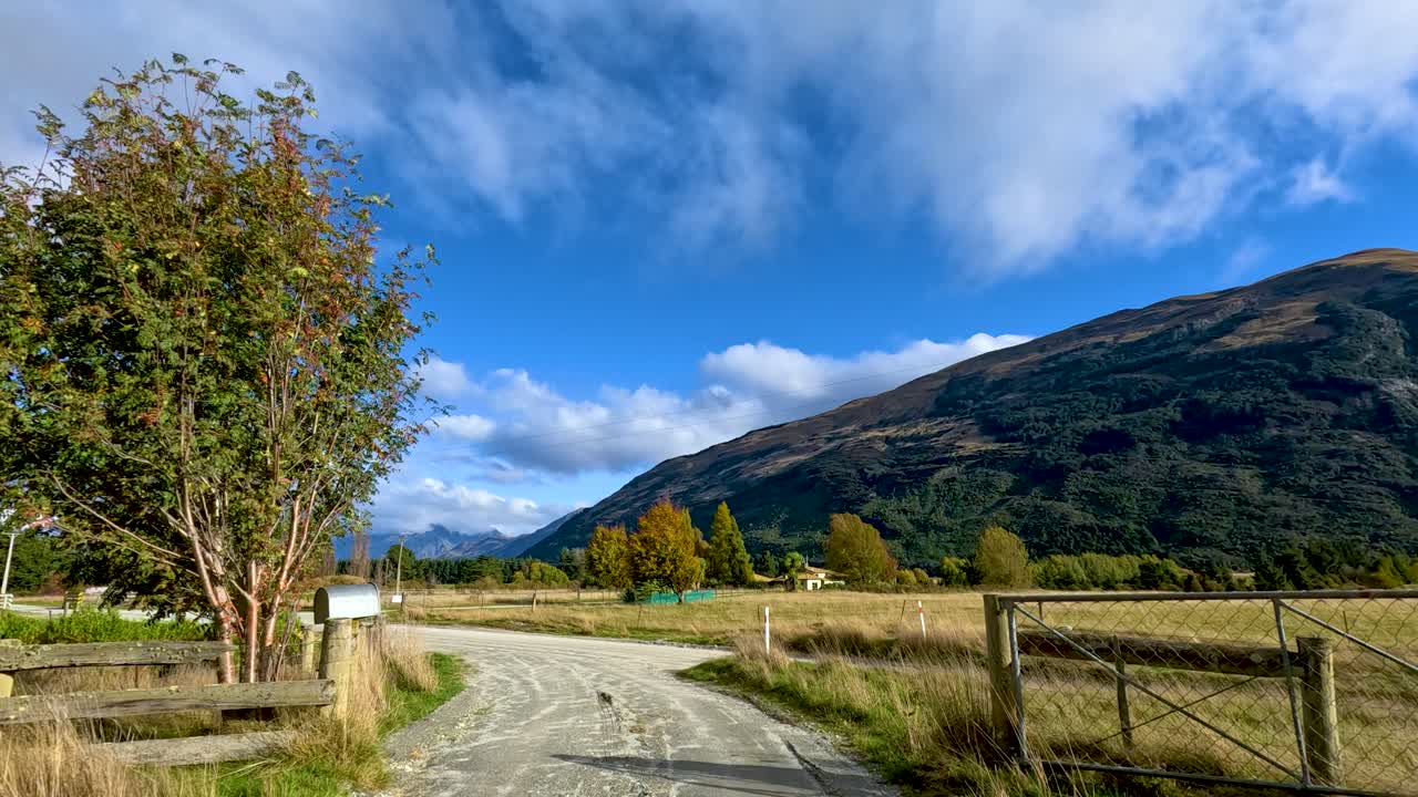 Vehicle drives along rural gravel road past fields and trees under bright blue sky