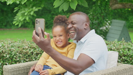 Grandfather-Pulling-Faces-With-Granddaughter-Taking-Selfie-On-Mobile-Phone-In-Garden-At-Home
