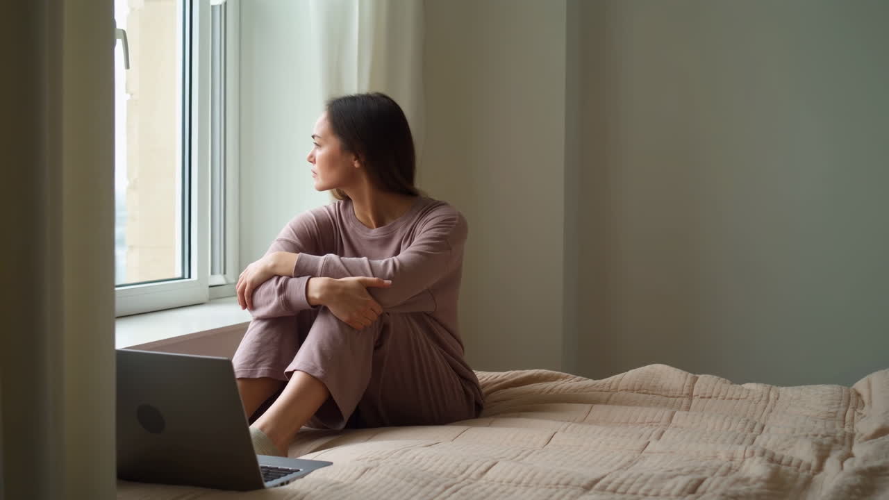 Donna seduta accanto alla finestra con laptop, luce soffusa mattutina sul viso, scrittrice freelance pensierosa sul letto, tonalità neutre.