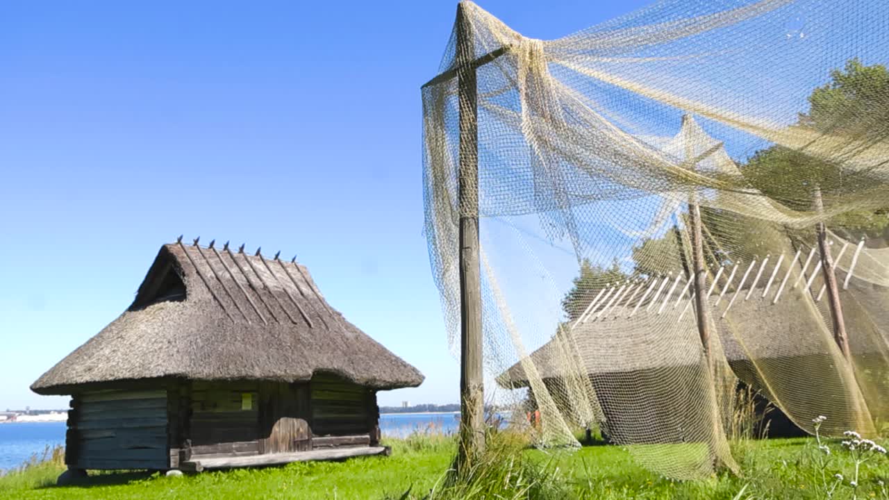 Gorgeous small fishing village with traditional woven fishing nets hanging on wooden poles and traditional old log buildings with thatched straw roof by sunny summer ocean shoreline seaside. Grassy
