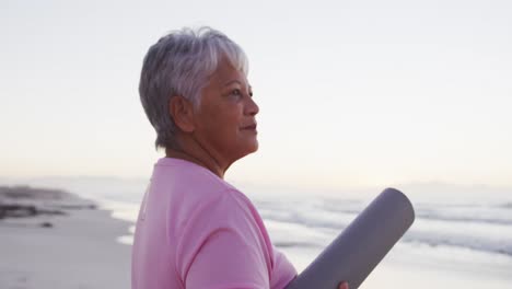 Eine-Afroamerikanische-Seniorin-Hält-Eine-Yogamatte-Und-Blickt-Auf-Die-Wellen,-Während-Sie-Am-Strand-Steht