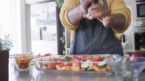 Midsection of african american woman in apron seasoning vegetables in kitchen, in slow motion