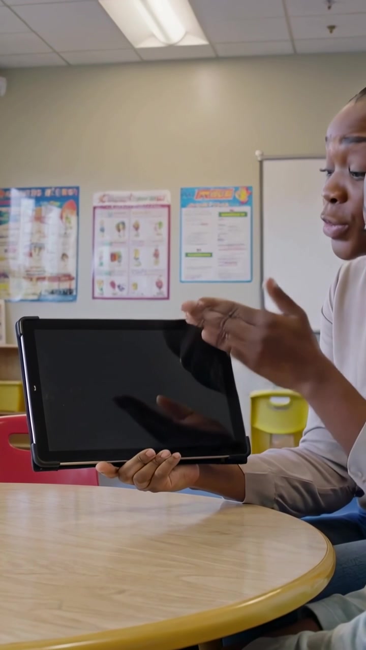 Vertical video: Placing tablet, woman in shirt showing screen to teach student in school