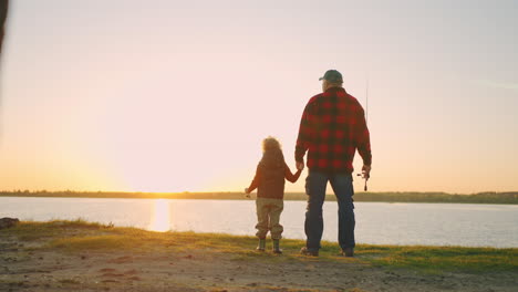 grandfather and little grandson are resting in nature standing on shore of river and admiring nature in sunset