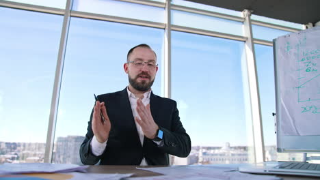 successful businessman leading a conference. male entrepreneur in glasses and costume sitting at the table and talking to someone in light office.