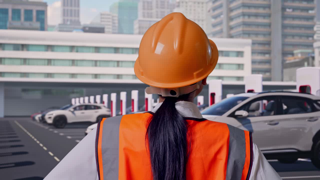 Primo piano da dietro di un'ingegnera con casco di sicurezza che si guarda intorno mentre è in piedi con le mani sui fianchi, veicoli elettrici in ricarica presso una moderna stazione di ricarica cittadina.