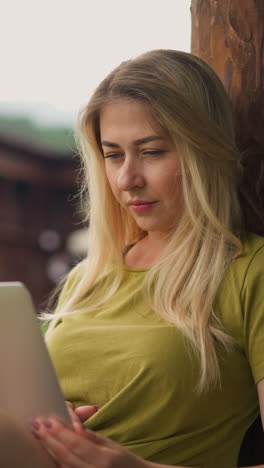 attractive lady with loose hair reads news in social media on laptop computer perching on restaurant terrace thick railing on rainy day slow motion