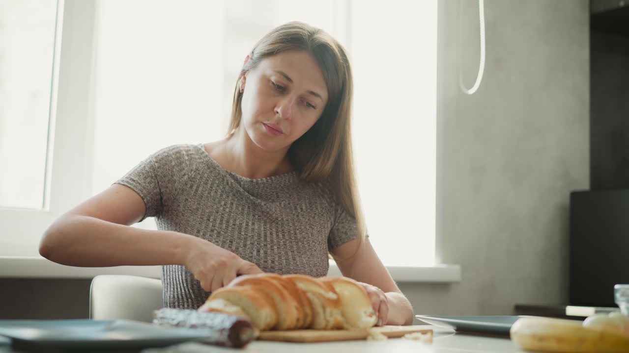 Medium shot of female confectioner seated by window slicing loaf of bread into equal slices on wooden board on table beside egg banana and plates under bright natural morning light