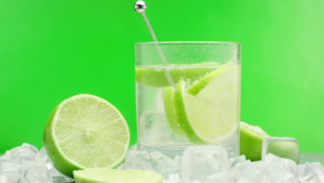 Slow-motion-product-video-of-a-refreshing-gin-tonic,-alcoholic-summer-drink-with-green-lemons-and-ice-blocks,-isolated-on-a-green-background,-rotating-on-a-turntable