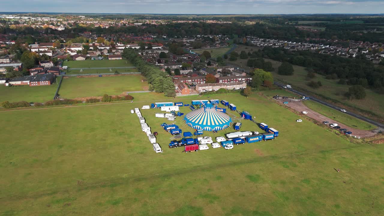An aerial view of a large field with a blue and white striped circus surrounded by vehicles, trucks, and trailers. The tent is set up in an open green space near a residential area