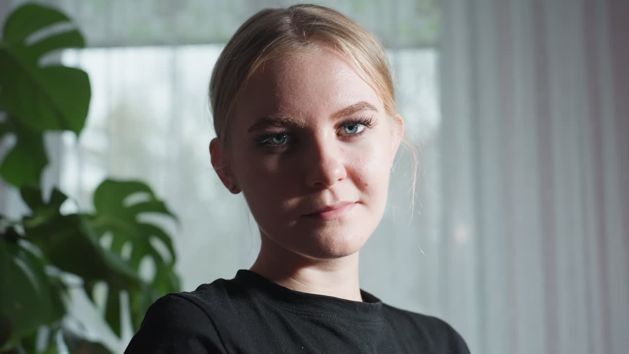 Close up of massage therapist with blonde hair and black shirt calmly looking at camera, standing near large leafy indoor plant with soft natural light filtering through sheer curtain in background