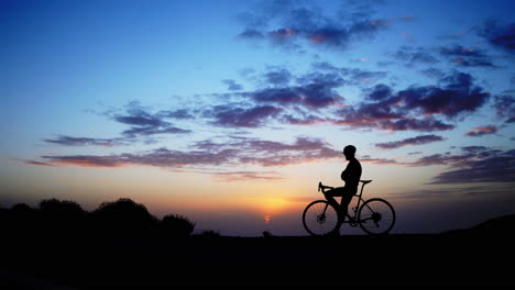 a man on a bicycle is positioned at the mountain's peak, observing the sunset. the camera glides using a steadicam for movement