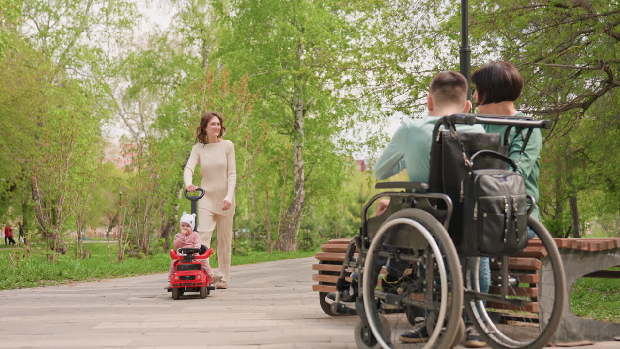Foreground Wheelchair And Caregiver Observing Child Approaching In Red Toy Car, Anticipation And Gentle Family Dynamic On Park Path, Candid Observation With Green Trees And Soft Light