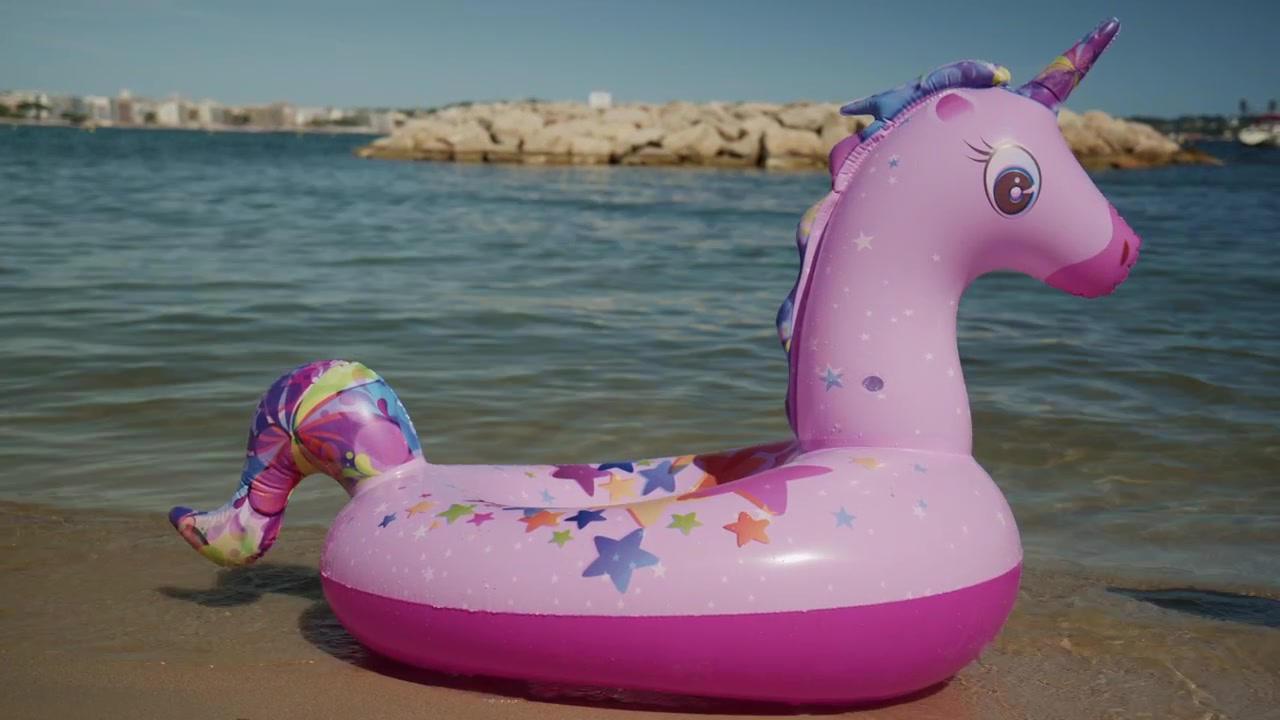 Phao bơi bơm hơi hình kỳ lân màu hồng nằm trên bờ cát của bãi biển Plage de la Gravette ở Antibes, Pháp, với làn nước Địa Trung Hải tĩnh lặng và đê chắn sóng đá ở phía sau