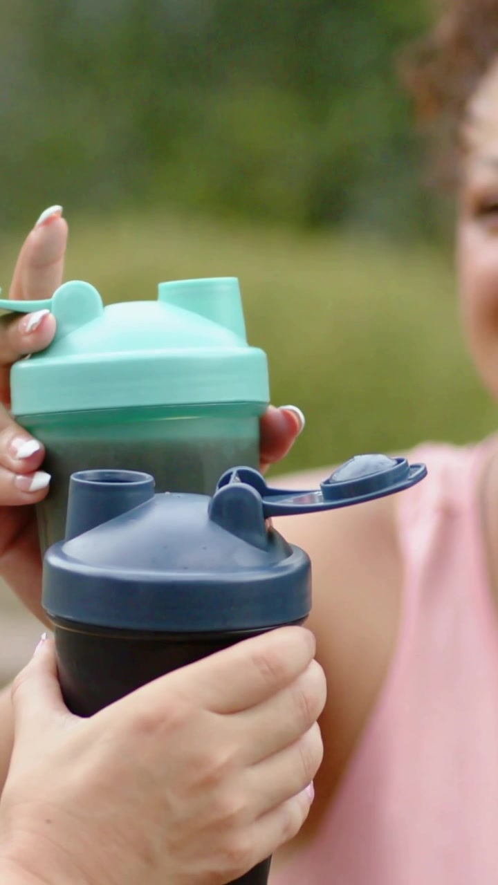 Video verticale: Una donna con un top rosa porta degli shaker verde acqua e blu scuro, apre i coperchi e brinda al parco.