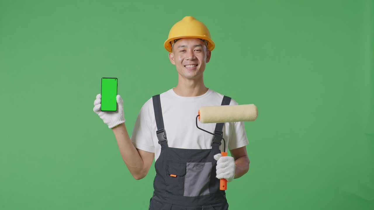 Asian Man Painter Wearing Safety Helmet Smiling And Showing Green Screen Smartphone To The Camera While Standing In The Green Screen Background Studio