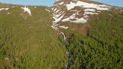 aerial view around a waterfall on a snowy mountain, sunny, summer day, in rotsund, troms, nordland, norway - orbit, drone shot