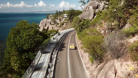 Vista-Aérea-De-Una-Mujer-Joven-Conduciendo-Un-Buggy-De-Playa-Clásico-Descapotable-Abierto-A-Lo-Largo-De-La-Carretera-Costera-Del-Acantilado-Junto-A-Aguas-Tropicales-Cristalinas