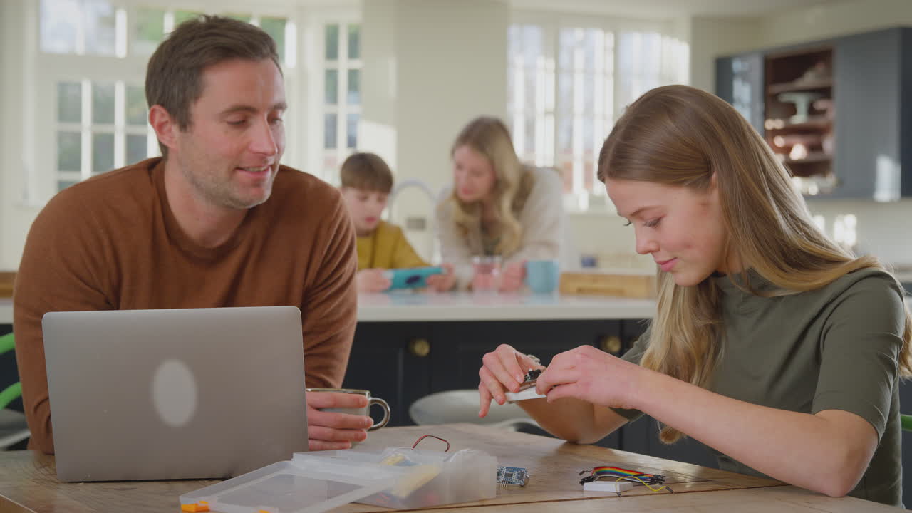 padre ayudando a su hija adolescente con un proyecto de electrónica sentado en la mesa de la cocina en casa con una computadora portátil - filmado en cámara lenta