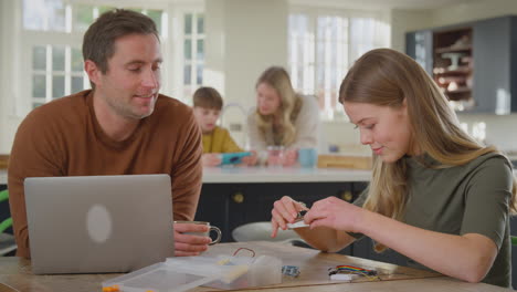 Padre-Ayudando-A-Su-Hija-Adolescente-Con-Un-Proyecto-Electrónico-Sentado-En-La-Mesa-De-La-Cocina-En-Casa-Con-Una-Computadora-Portátil---Filmado-En-Cámara-Lenta