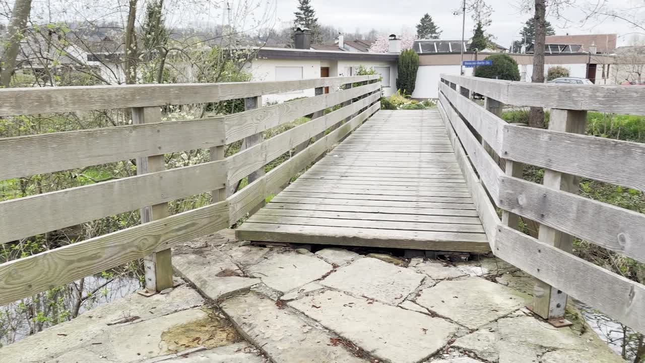 A small wooden bridge leading to a residential area; a river flows beneath it, panning shot