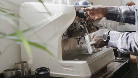 hands of african american male barista foaming milk at machine in coffee shop, slow motion