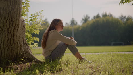 side view of young woman seated thoughtfully on grassy ground with sunlight illuminating her face, she rests her hand on folded leg, surrounded by nature, with stadium