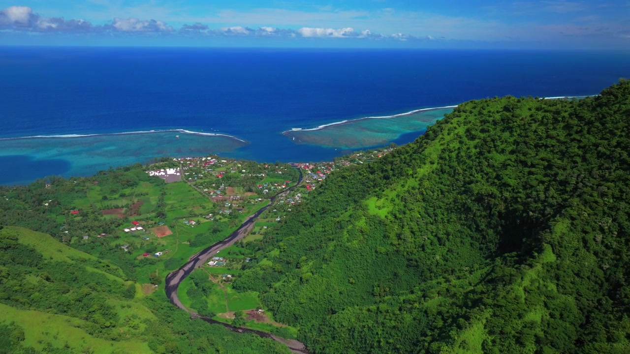 Veduta aerea di una soleggiata mattina nel villaggio di Teahupo'o, Fine della Strada, Tahiti, Polinesia Francese. Edifici del quartiere ripresi da drone, onda di Teahupo'o, laguna per il surf, barriera corallina di Passe Havea, valle del fiume Vaiau in primo piano.