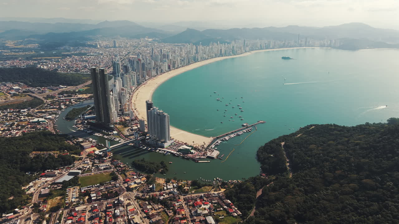 Drone view of Balneário Camboriú showing the bay, tall buildings along the beach, surrounding vegetation, port below, boats arriving, anchored vessels, and distant mountains