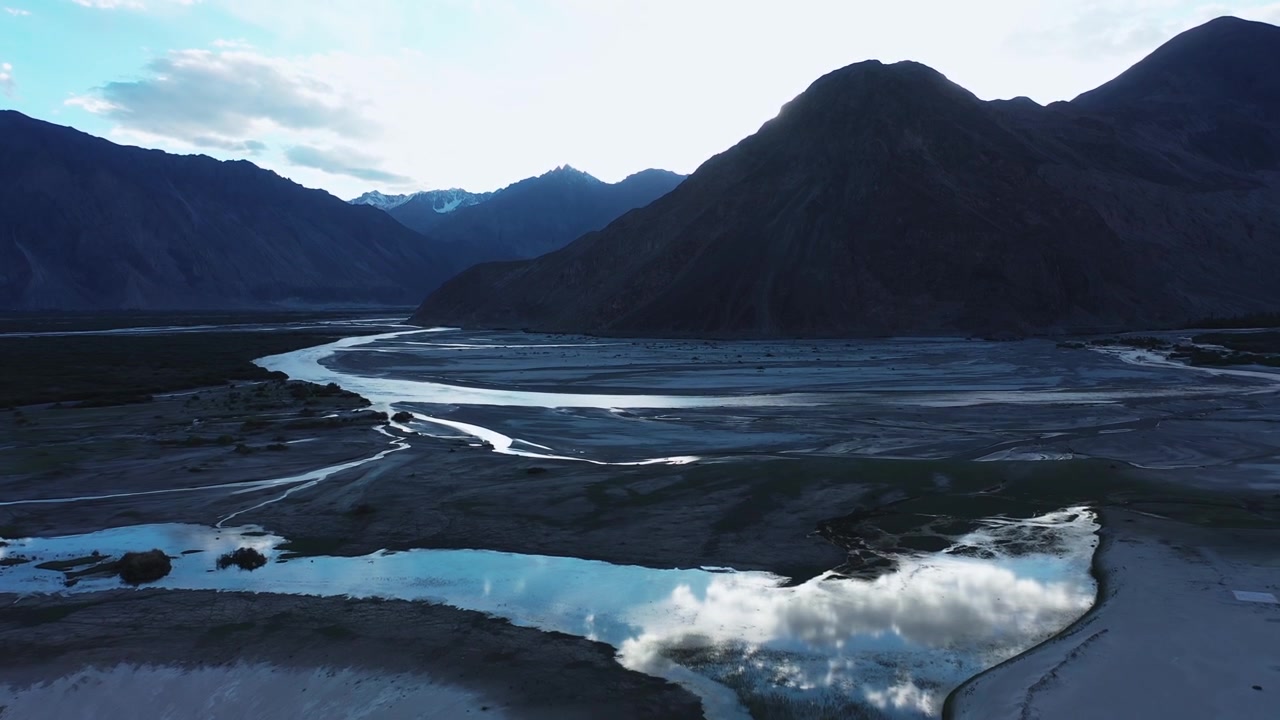 Nubra-dalens flettede flod snor sig gennem mørke sandklitter under Himalaya-toppe i skumringen. Droneoptagelse fanger reflekterende vandpytter og bjergsilhuetter i aftenlyset.