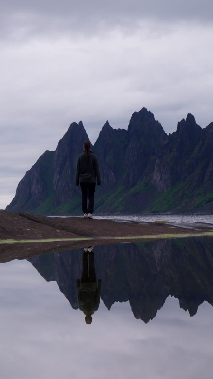 Dramatic cliffs, Devil’s Teeth, rise from sea in Senja, Norway under overcast sky, woman walks out onto sandbank, perfect reflection