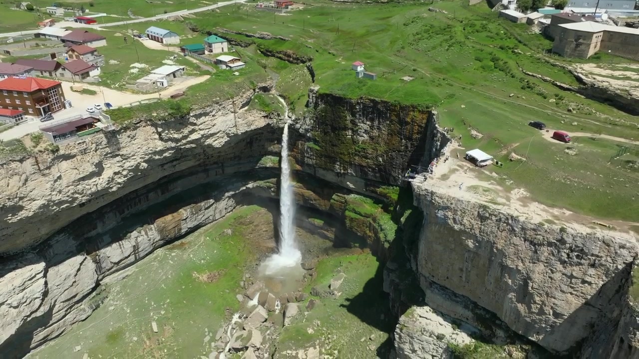 Rekaman udara Air Terjun Tobot yang terjun ke ngarai dalam di samping desa di Dataran Tinggi Khunzakh, Dagestan, Rusia, dengan ladang hijau dan rumah-rumah di atasnya.