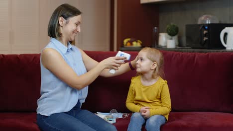 Mother-measures-temperature-with-contactless-digital-thermometer-of-her-cute-daughter-child-at-home