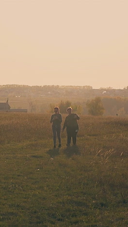 joyful young women walk dancing on meadow with dry grass against distant houses in early autumn morning slow motion