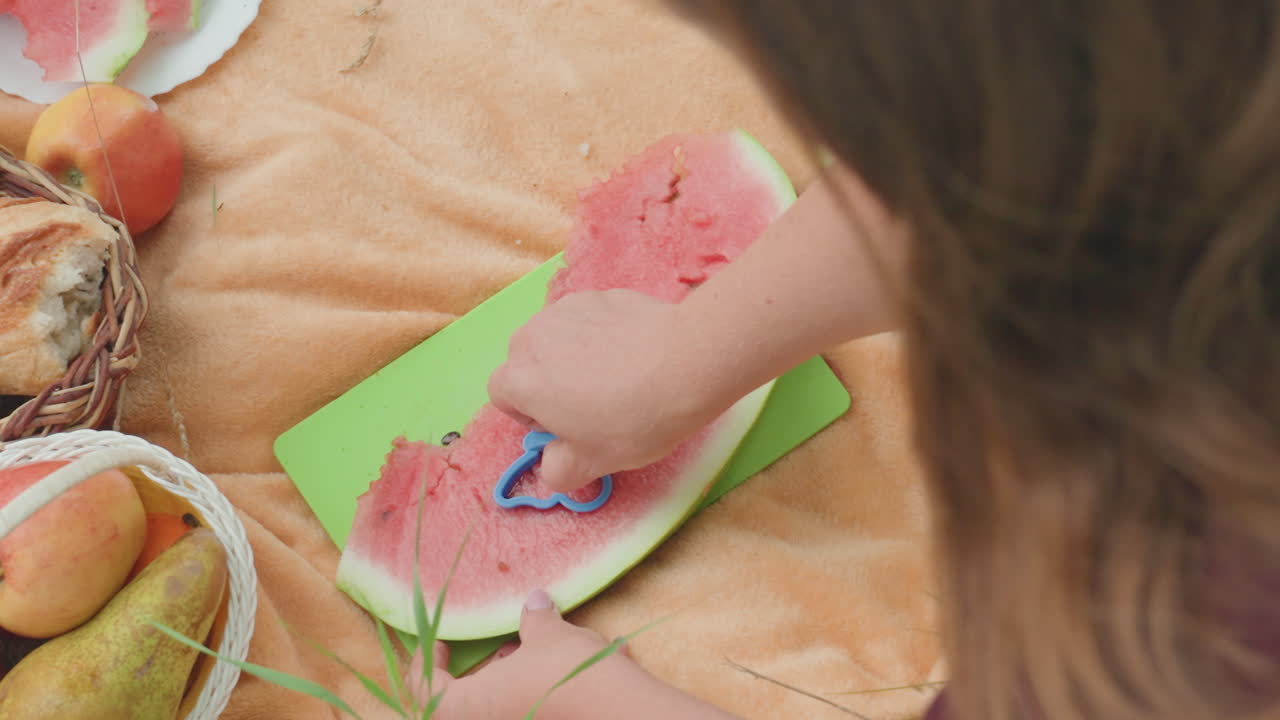 Niño tallando una sandía sobre una manta de pícnic con una tabla de cortar verde, un cortador de galletas, una cesta de melocotones y peras, una toalla color canela, primer plano de unas manos dando forma al melón para convertirlo en un tentempié divertido bajo la cálida luz del sol.