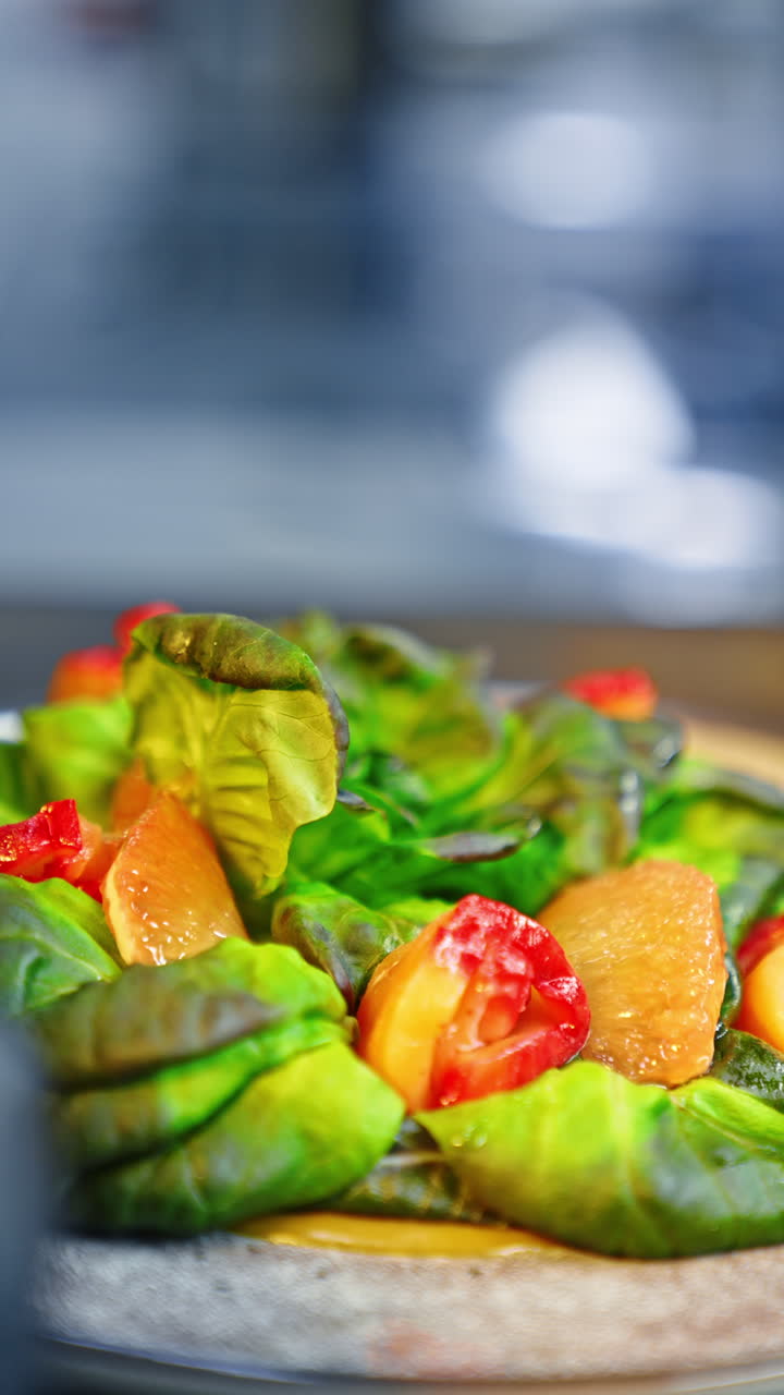 Fresh salad leaves and vegetables are on the plate. Cook's hands in black latex gloves put the pieces of tomato and grapefruit on the dish. Close up. Vertical video.