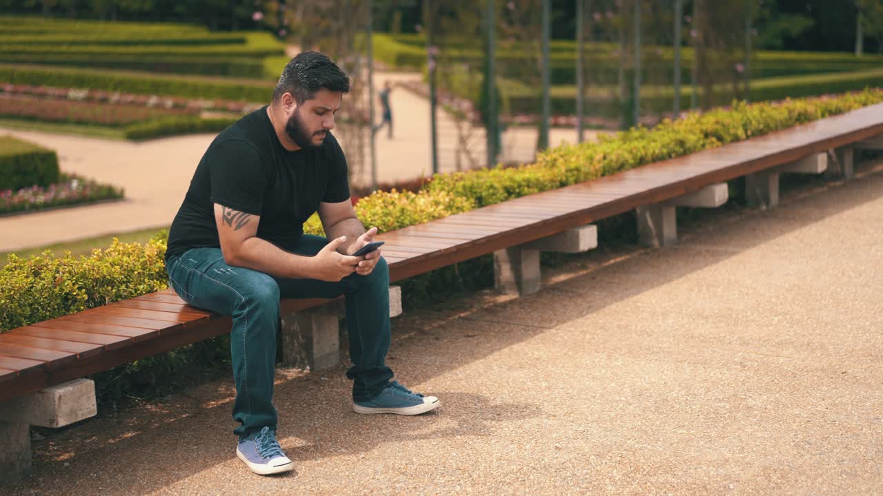 un joven que se levanta y envía mensajes de texto a su teléfono celular mientras está sentado en un banco largo en el parque al aire libre en cámara lenta
