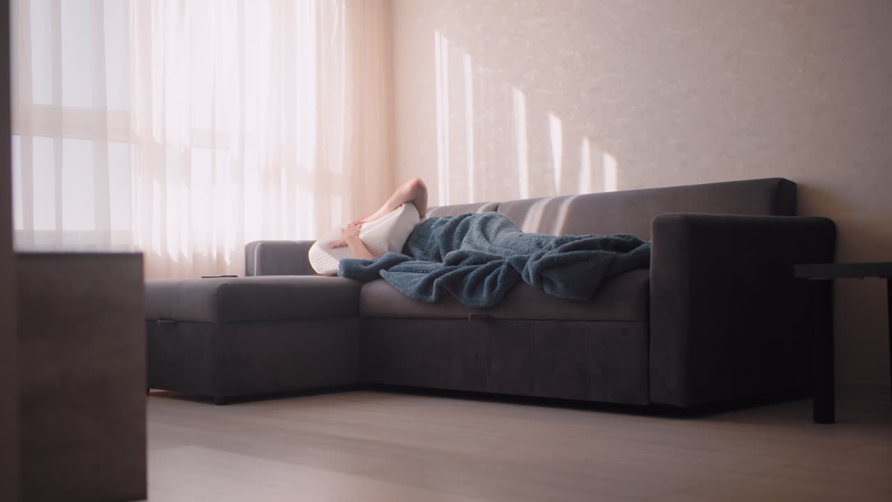 Young woman lying under blue blanket on grey couch in morning light, using pillow to cover head while avoiding sunlight, soft curtain shadows falling across wall