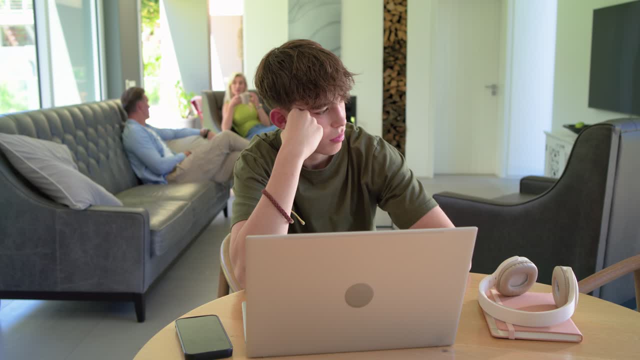 Family teenage boy typing on laptop, rubbing temples looking tired at home table as camera zooming