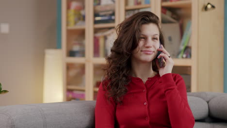 attractive caucasian woman in red cardigan on sofa smiles as she talks on phone