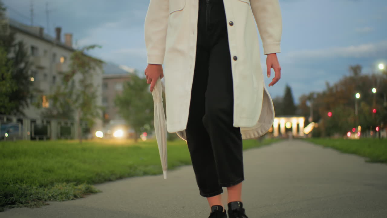 Woman wearing white coat strolling during evening holding umbrella with blurred moving cars, glowing streetlights, illuminated buildings and trees in vibrant cityscape under soft cloudy sky