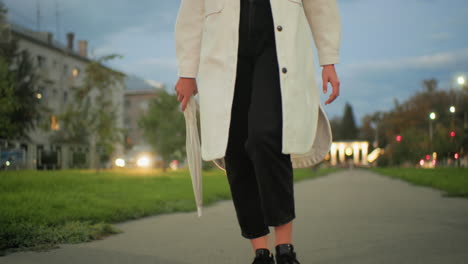 woman wearing white coat strolling during evening holding umbrella with blurred moving cars, glowing streetlights, illuminated buildings and trees in vibrant cityscape under soft cloudy sky